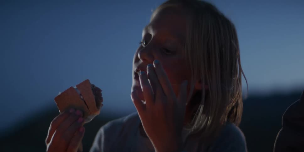 A young girl happily eats a smore outdoors at dusk her face illuminated by a warm glow