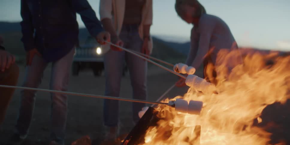 A low angle view shows a group of friends standing around a large bright campfire in the dark desert at dusk holding sticks with marshmallows over the flames to roast them