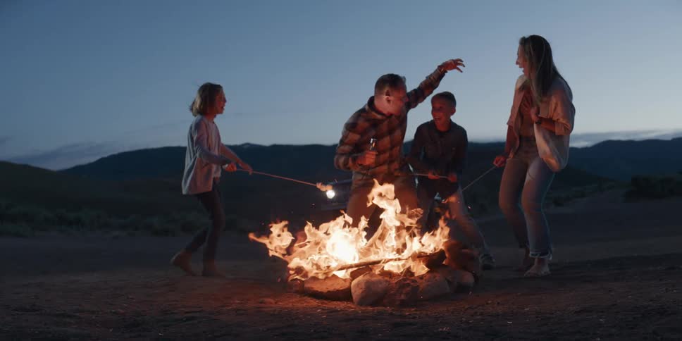 A family of four happily roasts marshmallows over a bright campfire at night in the desert with the man jokingly holding a flashlight under his face to tell a scary story