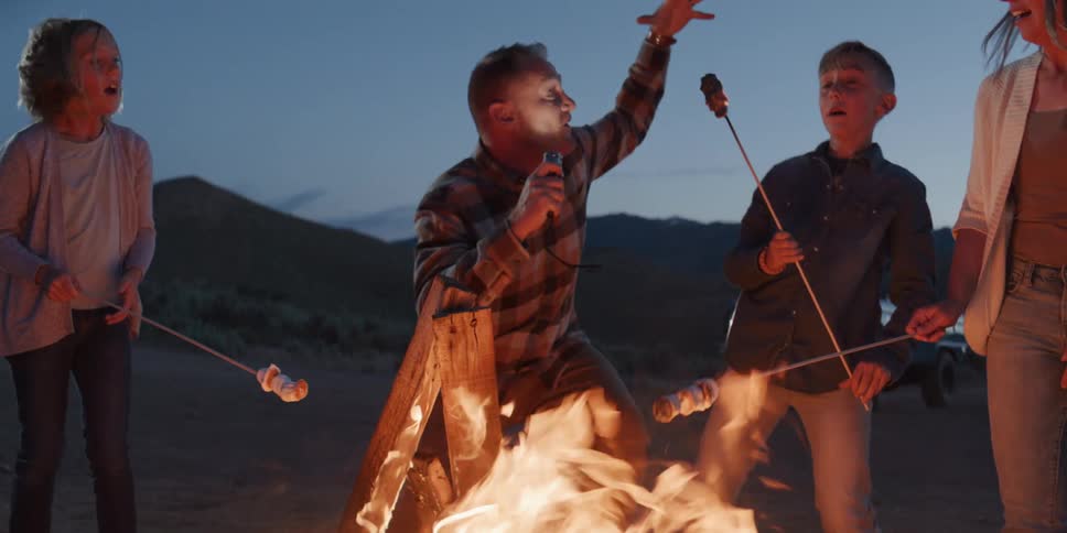 A group of people gathers around an open campfire at night the man in the center adds a log to the fire then raises his arm and then all people smile while holding sticks with food over the flames