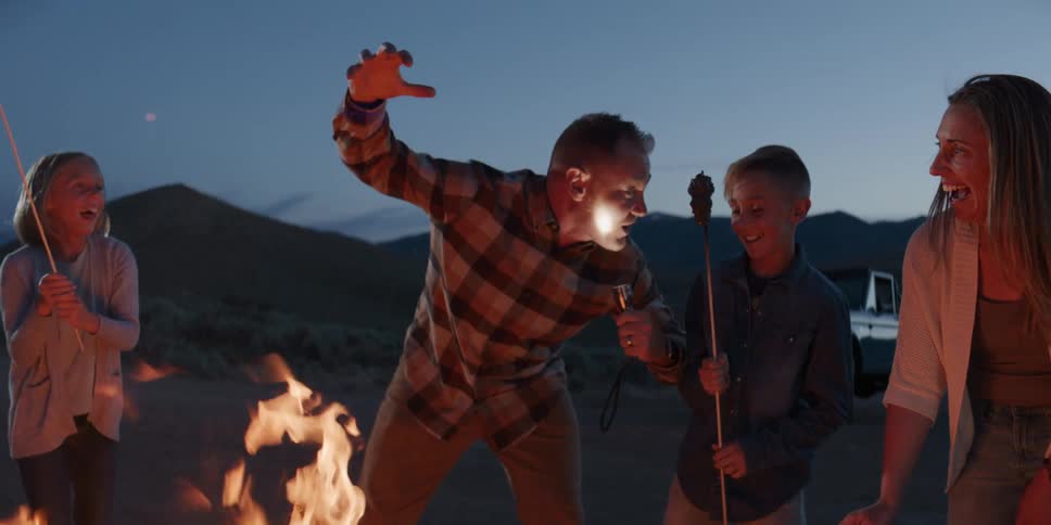Around a campfire at dusk a father playfully tells a spooky story with a flashlight to his laughing family as they roast marshmallows