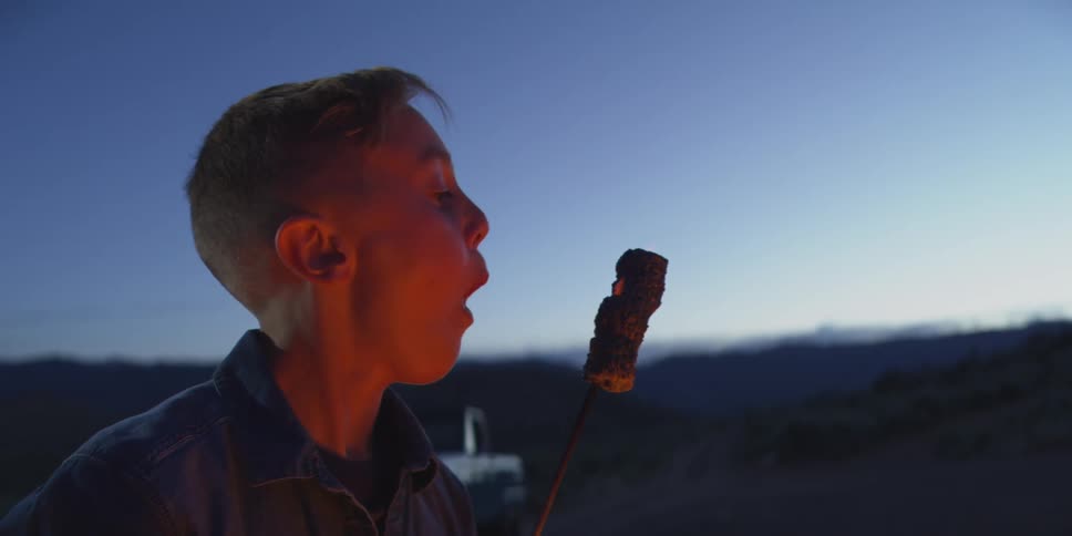 A close profile view shows a young man with a short haircut wearing a dark denim shirt roasting and eating a marshmallow over a glowing campfire at dusk in a dark open desert environment
