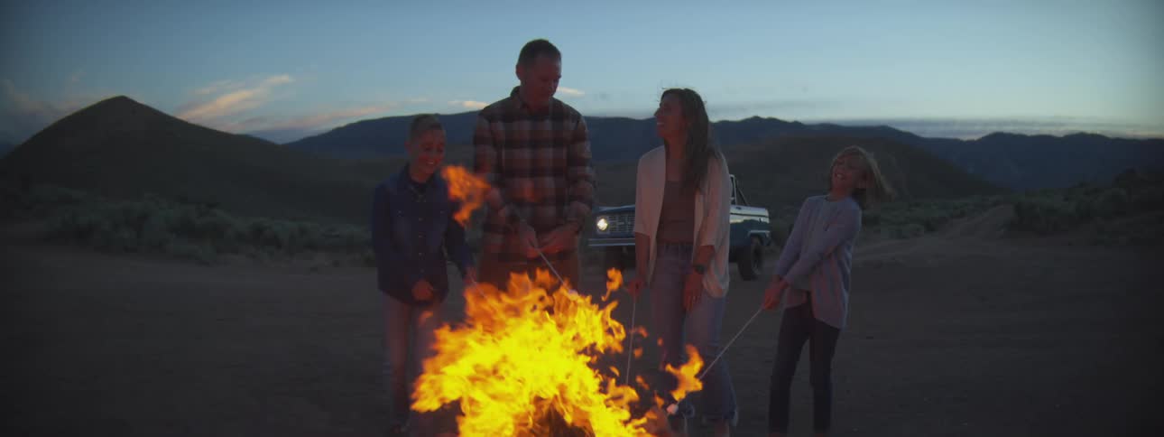 Family roasts marshmallows while standing around a campfire built in arid terrain after dusk