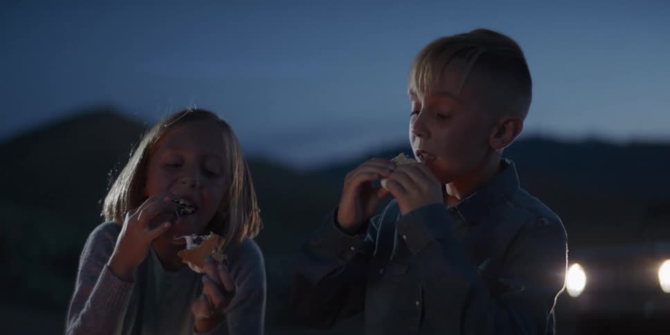 A close view shows a young child and a young girl eating messy smores with roasted marshmallows at a campsite at dusk illuminated by the warm glow of a nearby fire