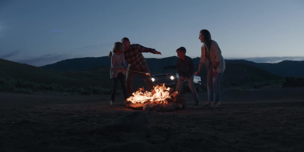 A smiling family of fourfather mother son and daughtergathers around a campfire joyfully roasting marshmallows together under the open sky