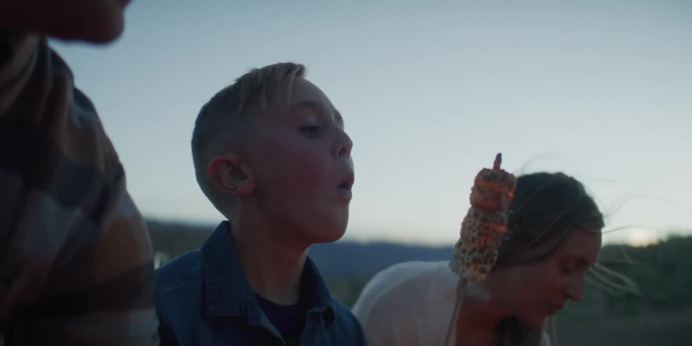 Family shares a moment outdoors with roasted corn in a mountainous setting at dusk