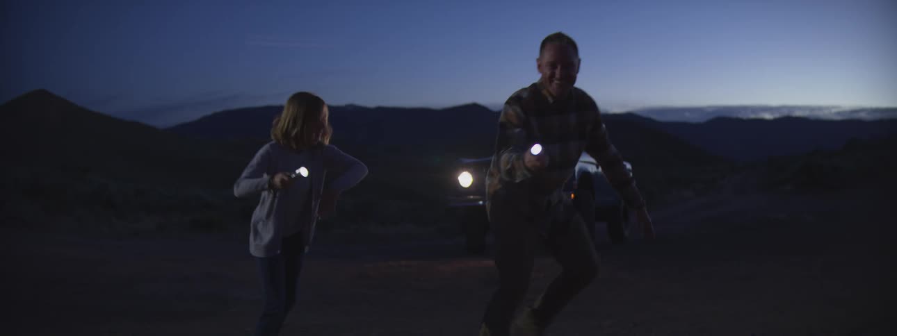 Family plays in the countryside between a roaring campfire and a parked vehicle at night