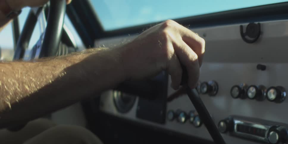 A man drives a car on a sunny day reaching down to shift the gear stick as sunlight streams through the windows