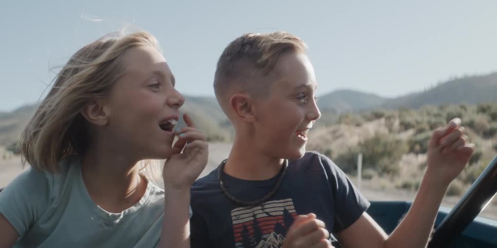 A closeup view shows a boy and girl in the back of a truck where the boy points excitedly into the distance and they both smile widely on a sunny day