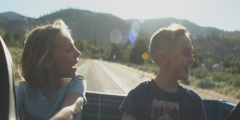 A boy and a girl sit in the back of an openroof car on a sunny day enjoying a road trip with the wind in their hair and smiles on their faces