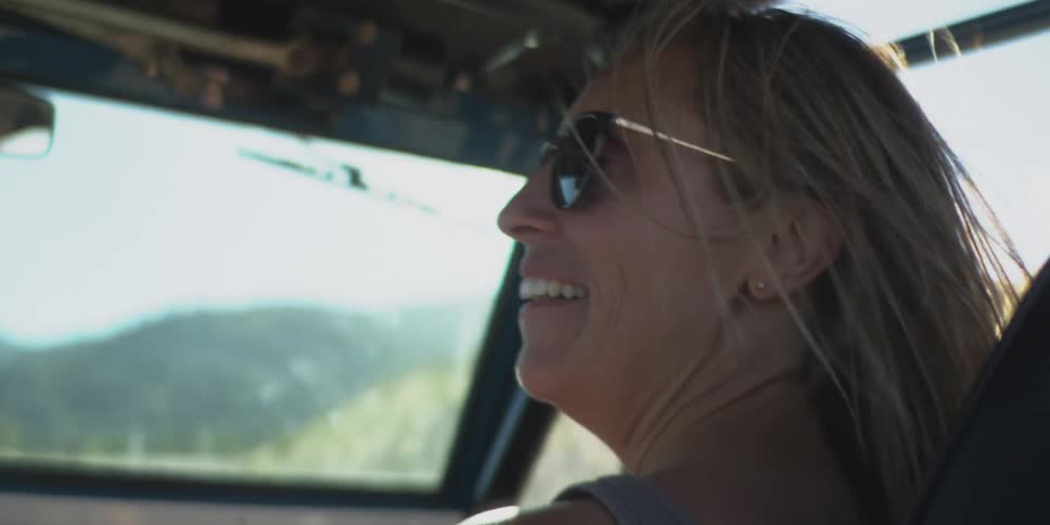 The video is a closeup of a woman driving a convertible or offroad vehicle with the top down on a sunny day her long hair is blowing in the wind and she is wearing sunglasses smiling as she looks out over the surrounding mountainous landscape