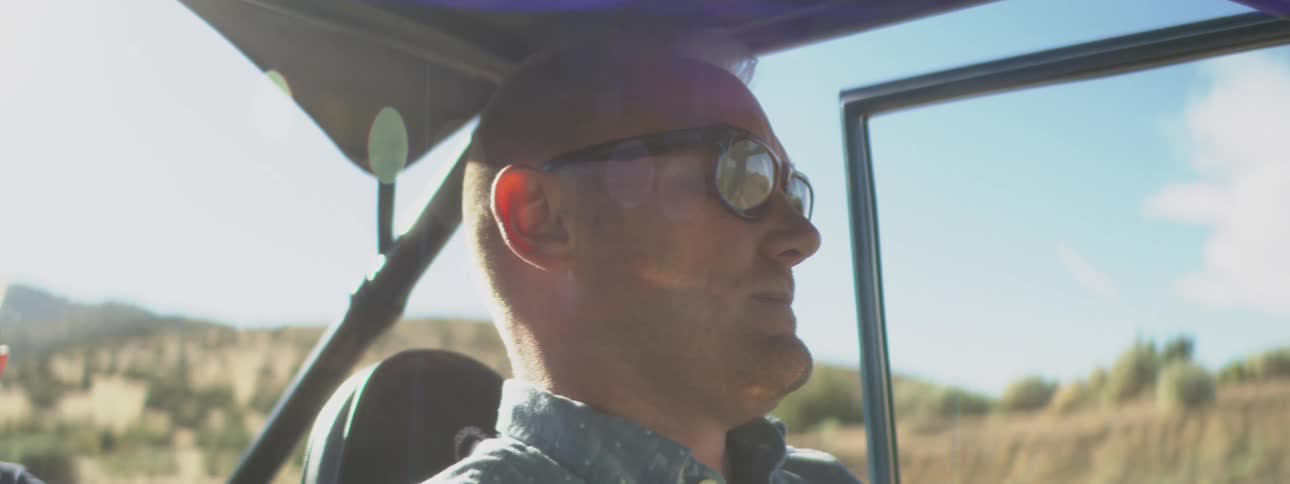 A closeup view of a mans face in profile shows him wearing sunglasses and riding in an open vehicle during bright daylight