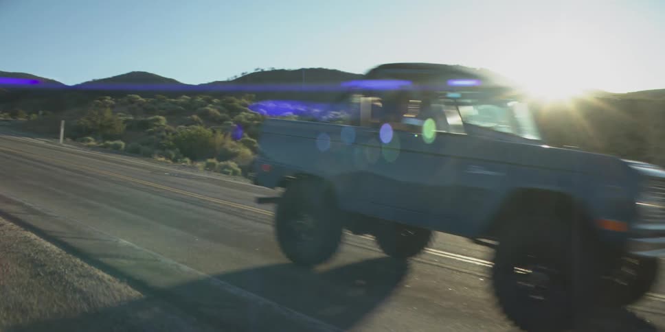 The video shows a dark blue suv or offroad vehicle driving toward the camera on a paved road in a mountainous desert landscape the sky is blue and clear and a bright sun flare is visible on the right side of the screen as the vehicle passes