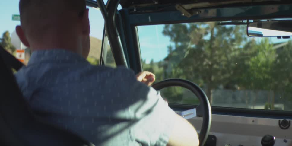 A view from the backseat shows a man in a blue patterned shirt driving a vintage offroad vehicle with a roll bar passing through a sunny suburban area with green trees and a hillside in the background