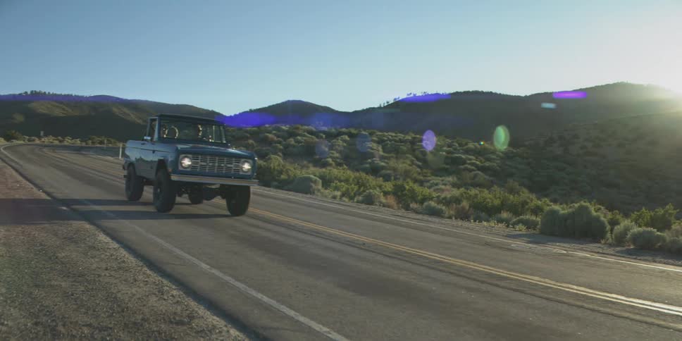 A low angle view shows a dark blue vintage offroad vehicle driving toward the camera on an open paved road surrounded by dry brush and hills with the bright sun creating a lens flare as it sets behind the hills