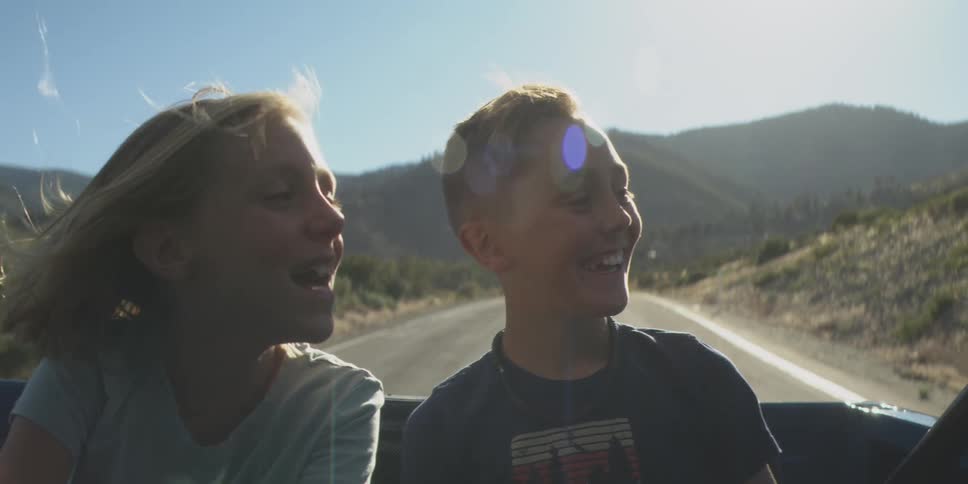 The video shows two children a boy and a girl riding in the back seat of a convertible or truck on a winding road they are facing each other laughing and enjoying the breeze with the sun in the background as they drive through a mountainous la