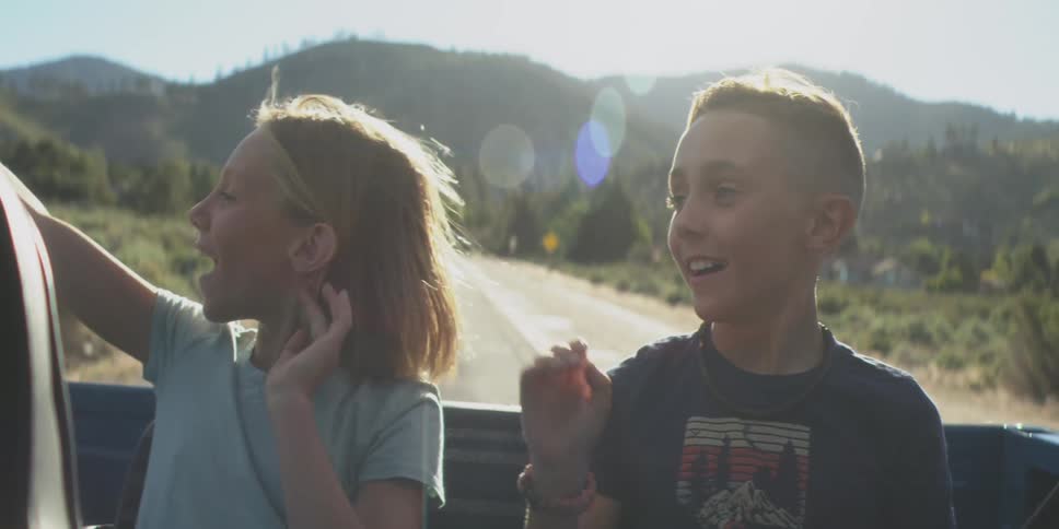 The video shows a young girl and a boy riding in the back of a truck or open vehicle on a sunny mountainous road they are both laughing with excitement and their hair is blowing dramatically in the wind as the truck moves quickly