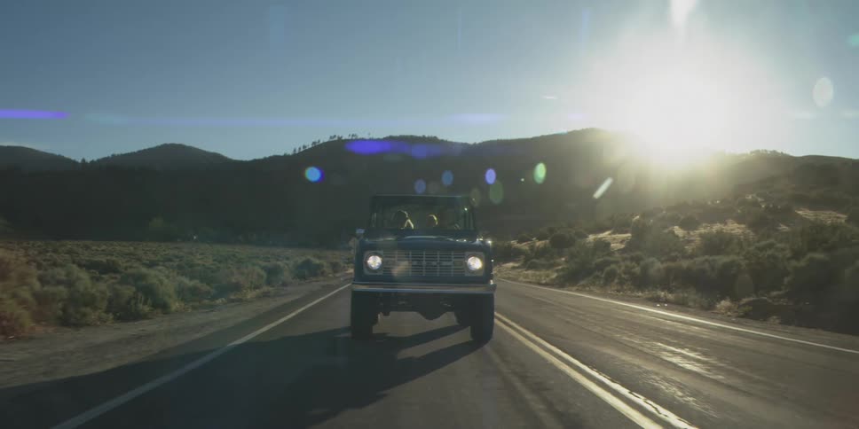 The video shows an old dark blue suv or offroad vehicle driving directly toward the camera on a winding paved road the scene is set in a mountainous desertlike landscape under a bright sun that creates a strong lens flare above the vehicl