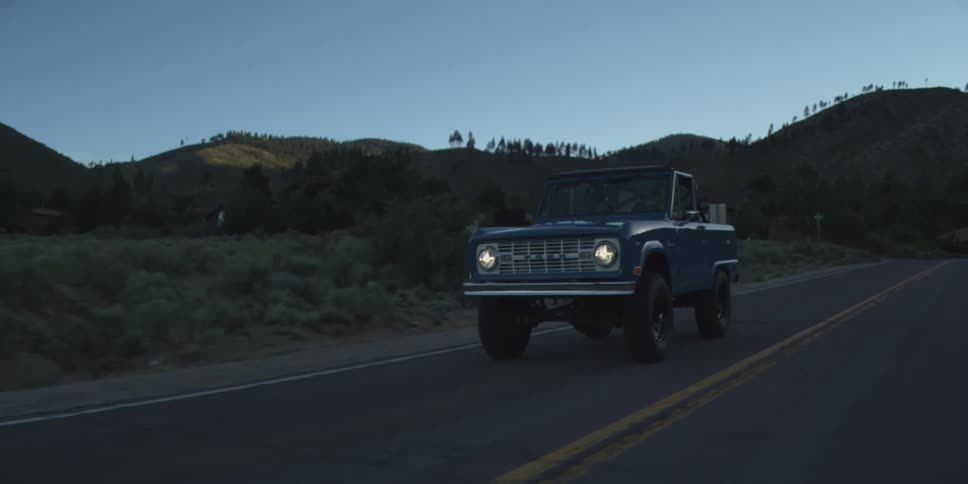 A car cruises along a deserted road at dawn its headlights cutting through the soft morning haze as the sky begins to glow with the first light of day