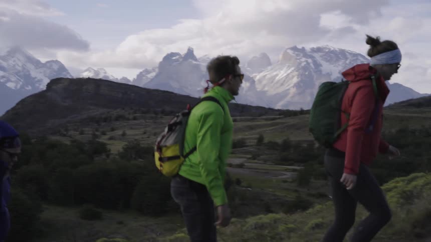 Three people hike up a hill with snow covered mountains in the distance