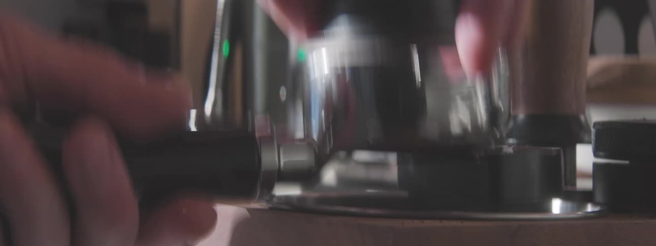 A close view shows a persons hands holding a black portafilter and using a tamper to compress ground coffee into the filter with a shiny espresso machine visible in the blurry background