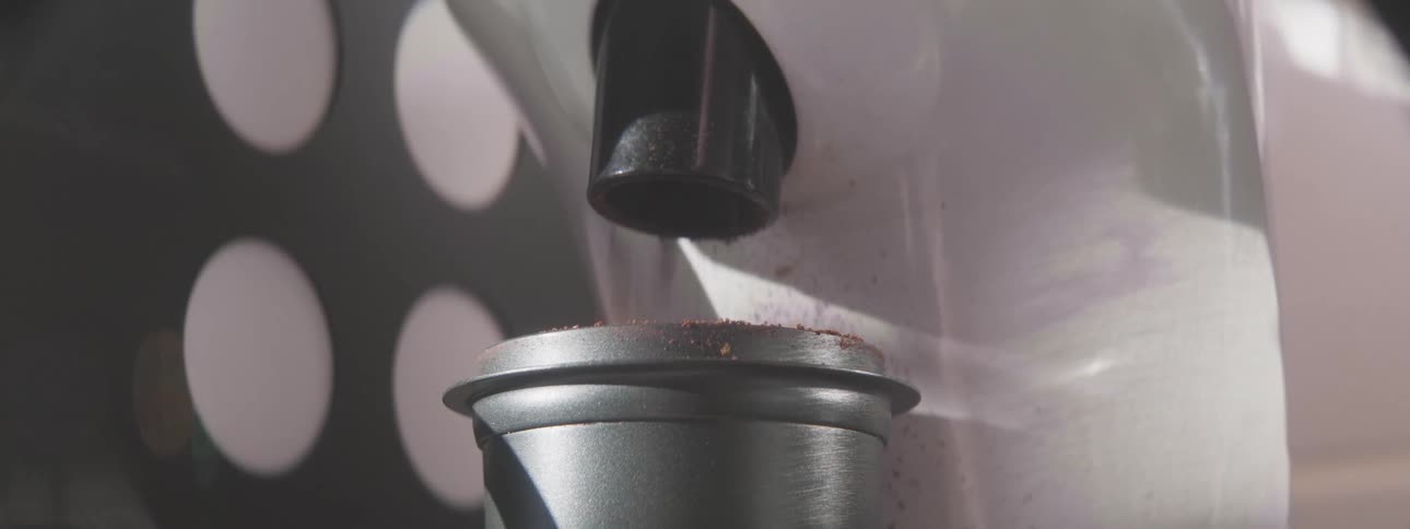 A close view of an espresso machine shows ground coffee pouring out of a black spout and accumulating in a metal portafilter cup against a bright patterned wall
