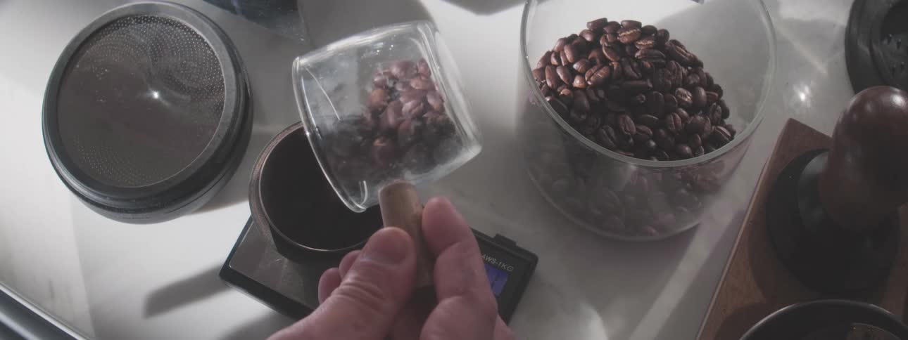 A persons hand places a metal dosing cup on a digital scale and then pours whole coffee beans from a small glass scoop into the cup next to a large jar of roasted coffee beans