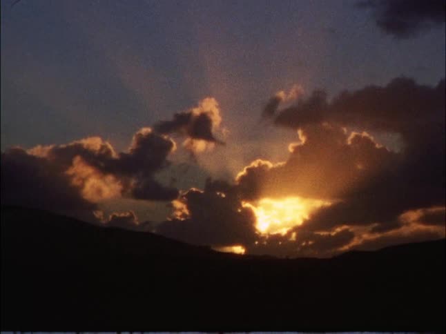 A dramatic sunset partially hidden by dark clouds over distant mountains