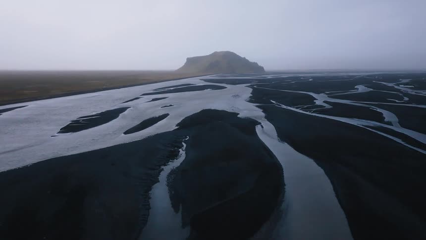 The video is an aerial view of a wide river splitting into multiple braided channels across a dark flat landscape in the distance there is a single prominent coneshaped mountain visible through the heavy mist or fog