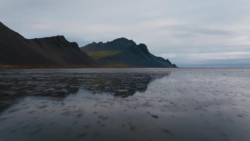 A dramatic wide shot shows dark jagged mountains reflected on a wide wet black sand beach under an overcast sky at dusk