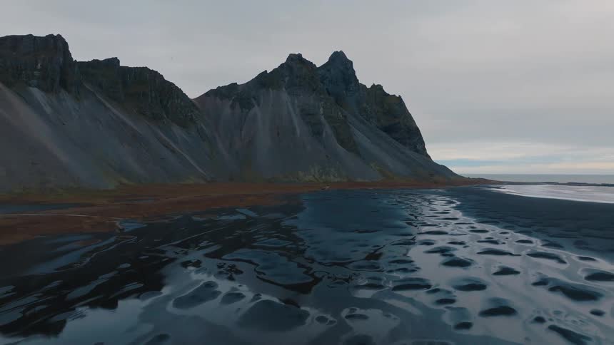 A wide shot shows a black sand beach and wet tidal pools leading up to the steep jagged peaks of a mountain range against an overcast sky