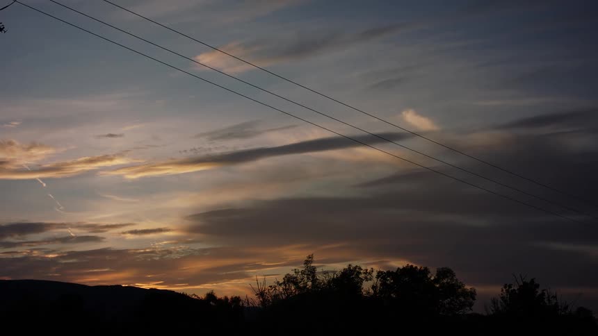 Power lines cross colorful sky above silhouettes of trees and mountains at dusk