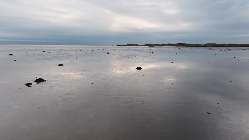 Dark rocks rest on reflective wet sand in flat coastal area under cloudy sky