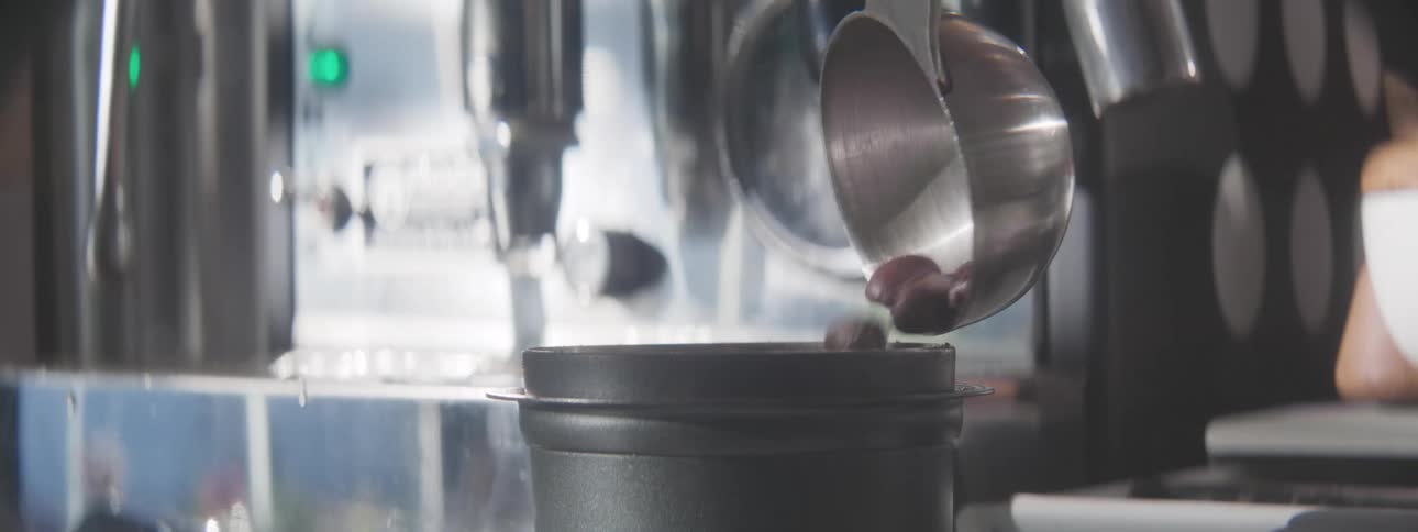 Closeup of coffee beans being poured from a metal scoop into a grinder in cafe