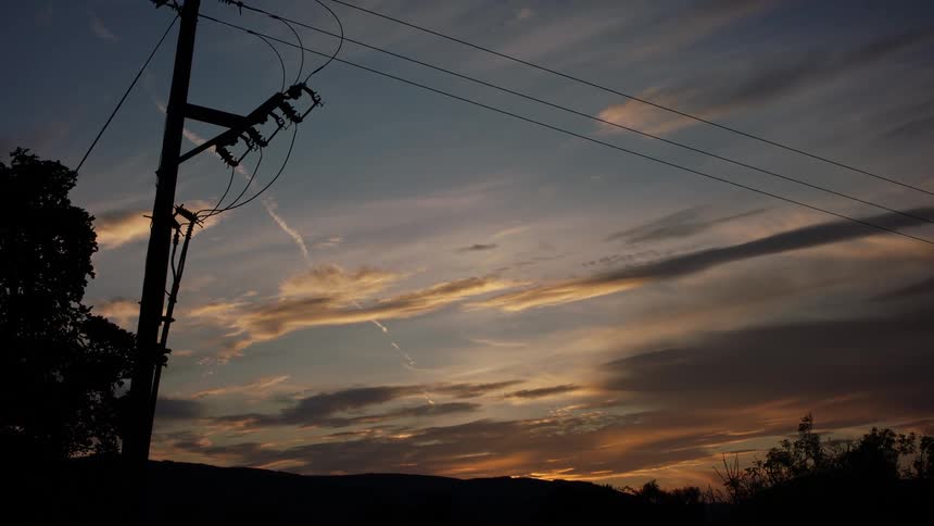 A wide shot shows a silhouette of an electrical utility pole and power lines running across a sky filled with clouds illuminated by the colors of a sunset or sunrise above a dark horizon