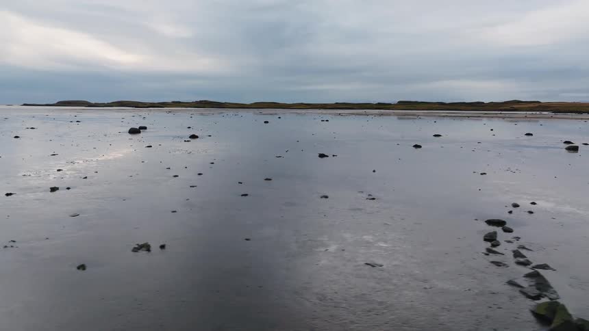 Flat coastal area with scattered rocks and water puddles under cloudy sky