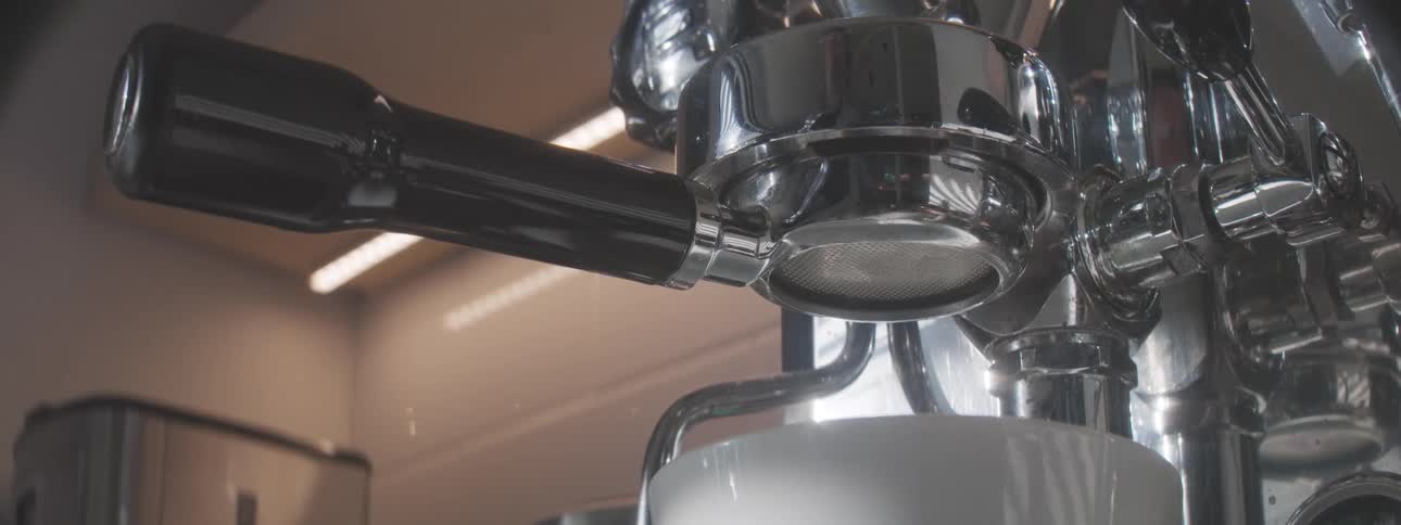 A close view shows a hand inserting a portafilter into the group head of a shiny chrome espresso machine then a white cup is placed below and rich espresso begins to pour into it