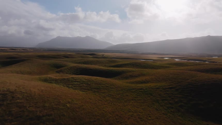 An aerial wide shot shows rolling brown and green grassy hills in the foreground with high dark mountains in the background under a dramatically cloudy and bright sky