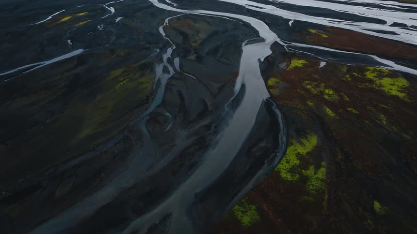 An aerial full view shows a braided river with multiple pale blue gray channels flowing through dark volcanic soil and patches of yellow green mossy tundra