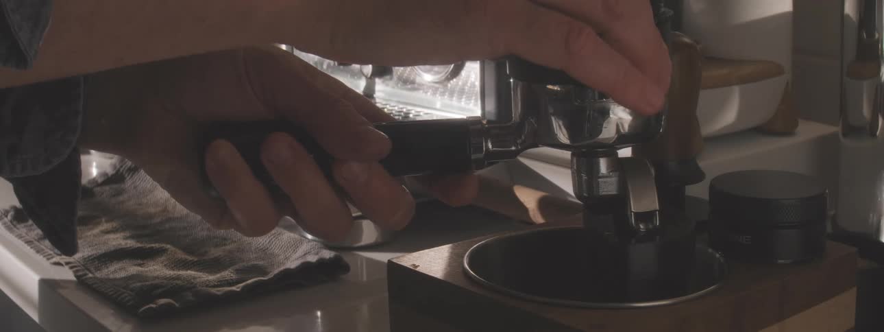 A closeup view shows a persons hands holding an espresso portafilter containing coffee grounds then inserting a tamper and pressing down on the grounds to prepare for brewing next to a chrome espresso machine