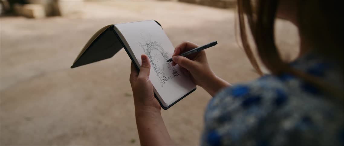 Close up of a young woman sketching in a drawing book while sitting in a corridor during daytime