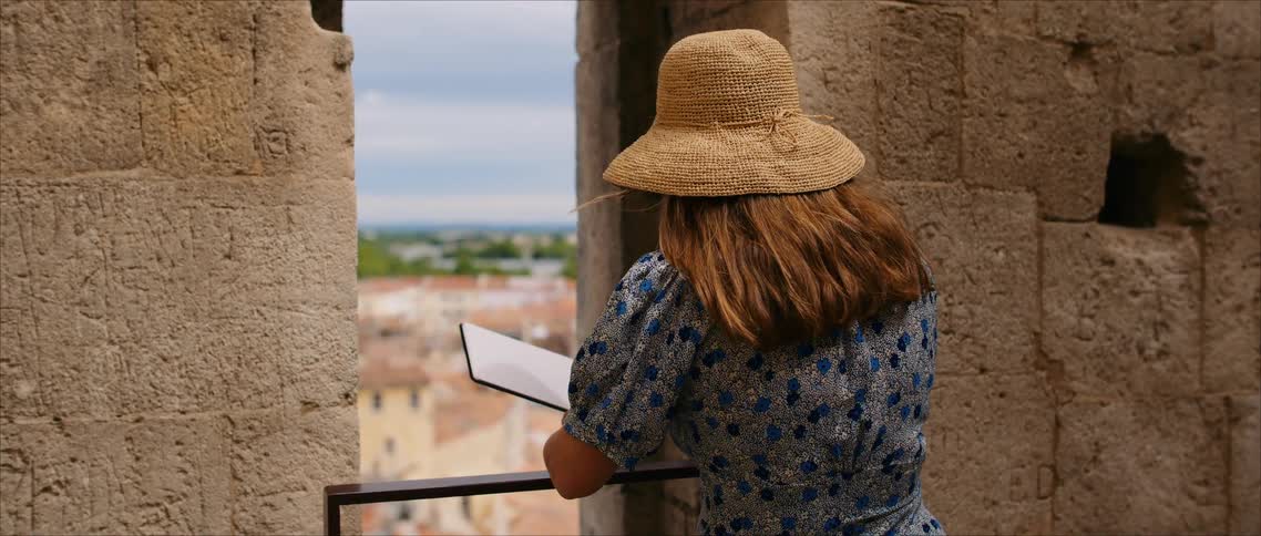 Woman wearing a straw hat sketches in a notebook while observing the cityscape