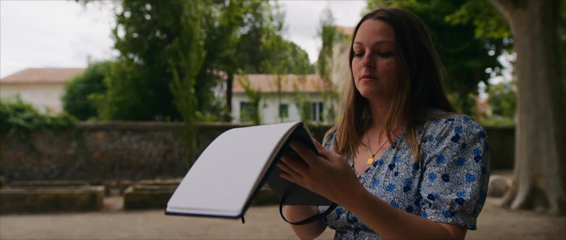 A young woman reads from a notepad outdoors at midday focused and relaxed as sunlight filters across the open space around her