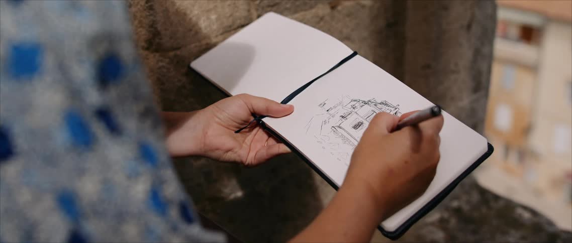 Person sketches architectural structure in white notebook in front of stone wall in urban setting