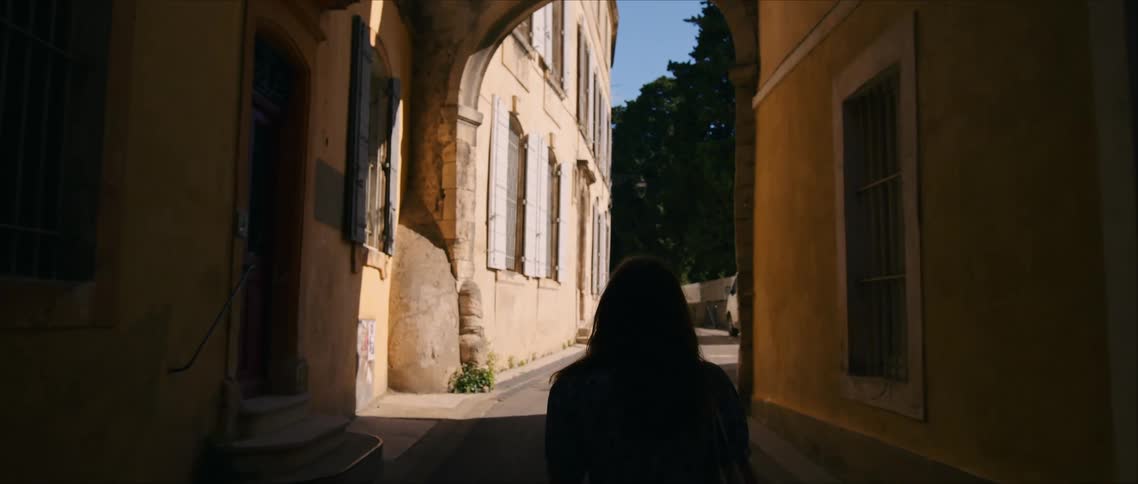 A woman walks through a narrow sunlit alley beneath an old stone arch toward a bright courtyard