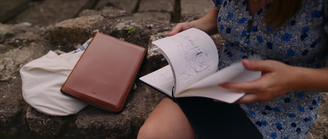 A person sits on rocky ground flipping through a sketchbook under daylight