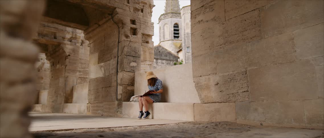 Person sits on stone bench facing historic structure with tower in background under clear sky