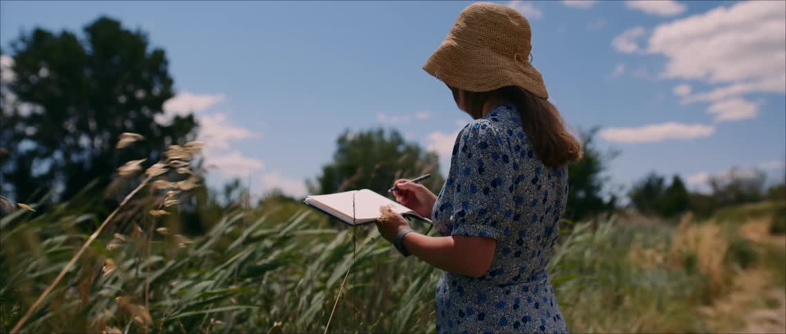 A woman with long brown hair wearing a blue patterned dress and a straw sun hat stands in a tall grassy field writing in a notebook on a bright sunny day