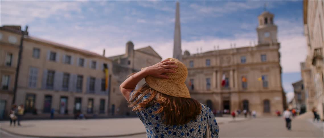 A woman in a blue patterned dress and a straw hat stands in a sunny european square with her hand touching the hat looking toward historic buildings and a tall spire