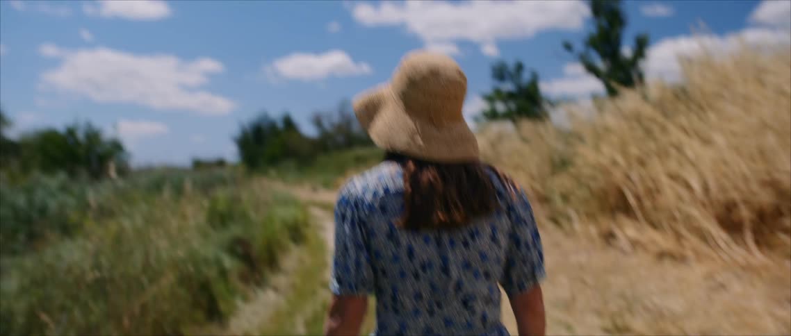Woman in floral dress and straw hat walks along path between dry grass and greenery under clear sky